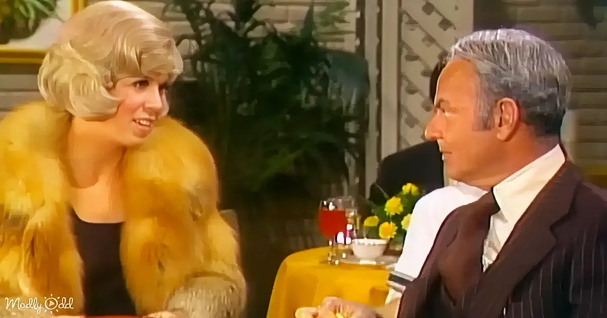 A woman in a yellow fur coat smiles at a man in a suit at a table with drinks.