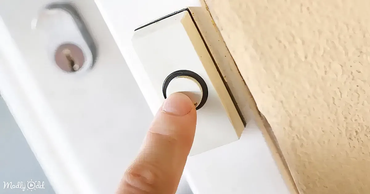 A finger presses a black button on a white door with a keyhole and textured wall.