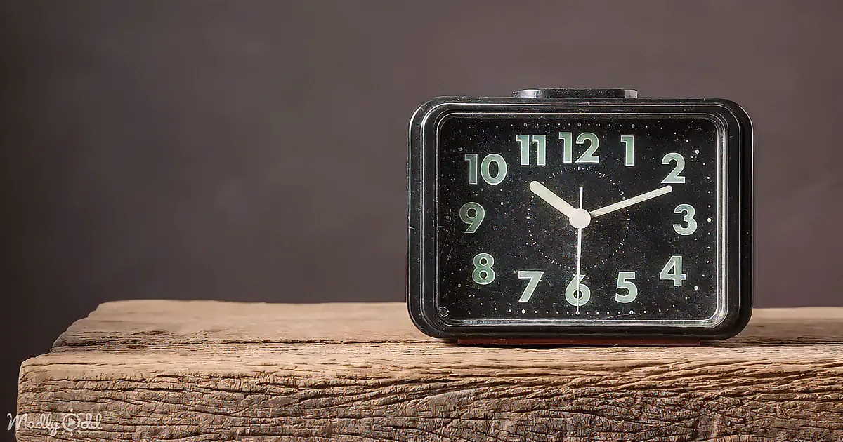A vintage black alarm clock with white numbers and hands sits on a wooden surface against a dark background.