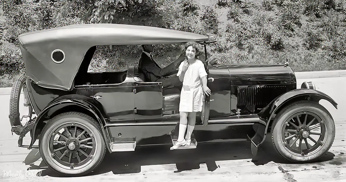 A young girl stands on the step of a vintage car, smiling, with a man inside the vehicle.