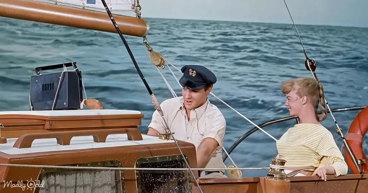 A man in a sailor's uniform and a woman in a striped shirt are on a sailboat, smiling and talking.