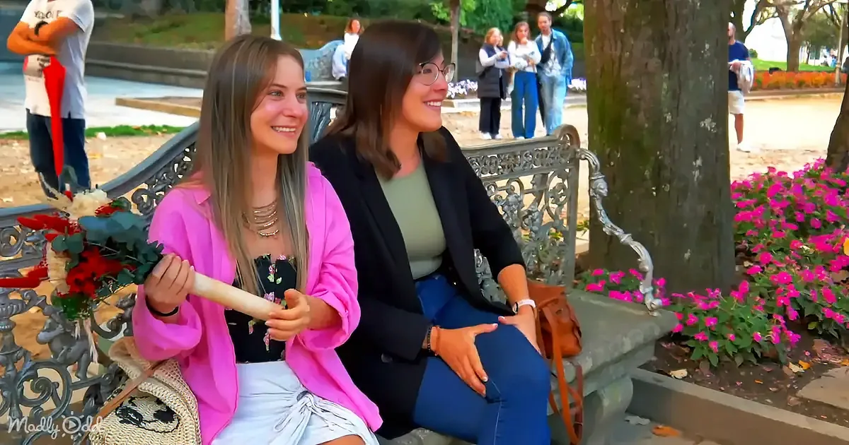 Two women sit on a bench in a park, smiling and holding a rolled-up document.