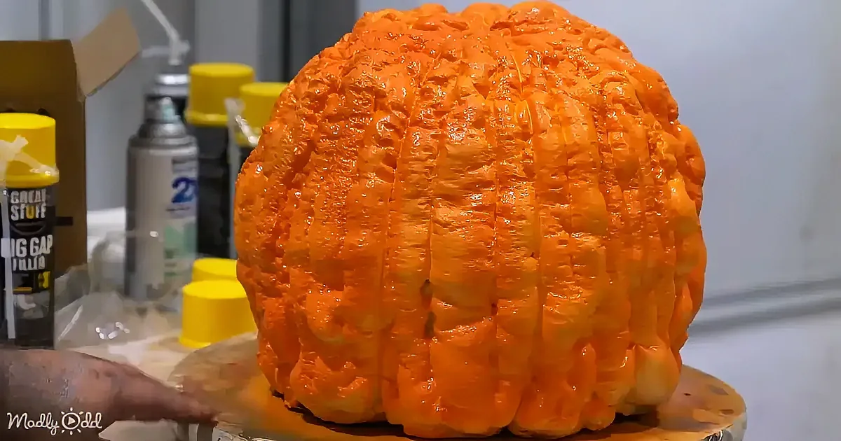 A large, textured orange pumpkin sits on a table with spray cans and other items in the background.