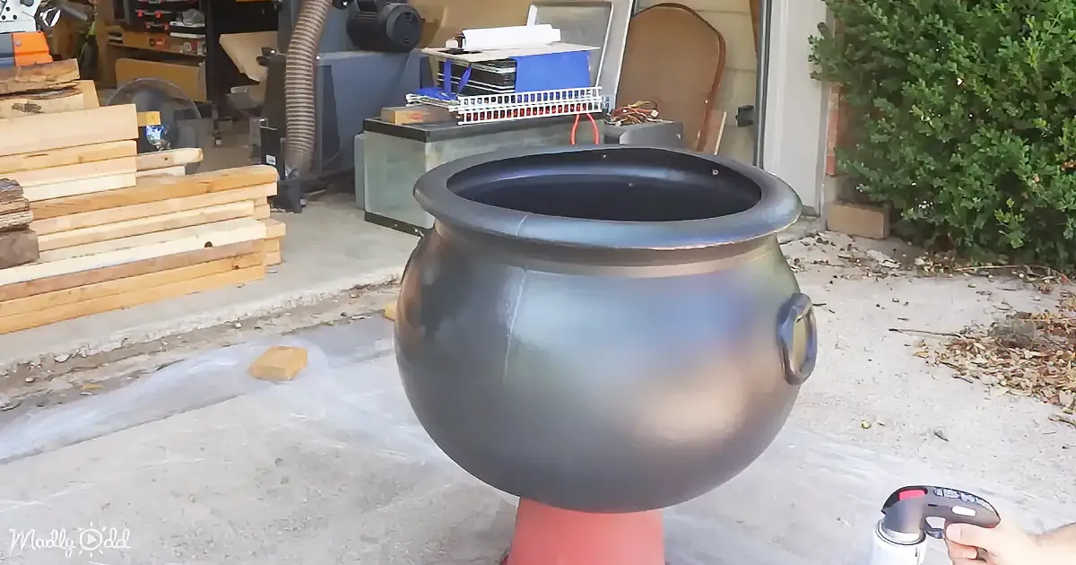 A large, shiny, black cauldron sits on a red stand in a workshop with tools and wood in the background.