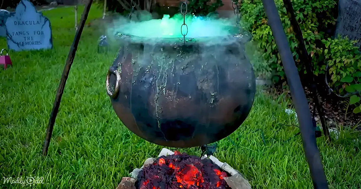 A steaming cauldron hangs over glowing embers, surrounded by Halloween decorations in a grassy yard.