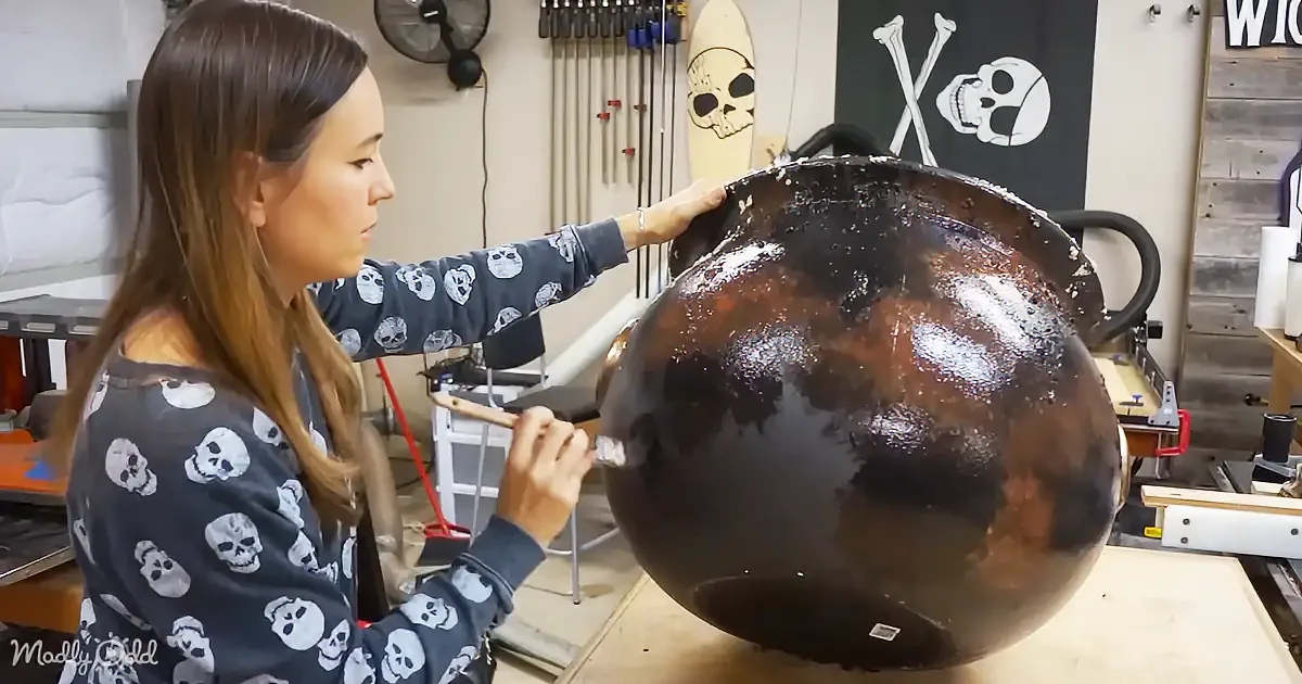 A woman in a skull-patterned shirt is painting a large, dark sphere in a workshop filled with tools and decor.