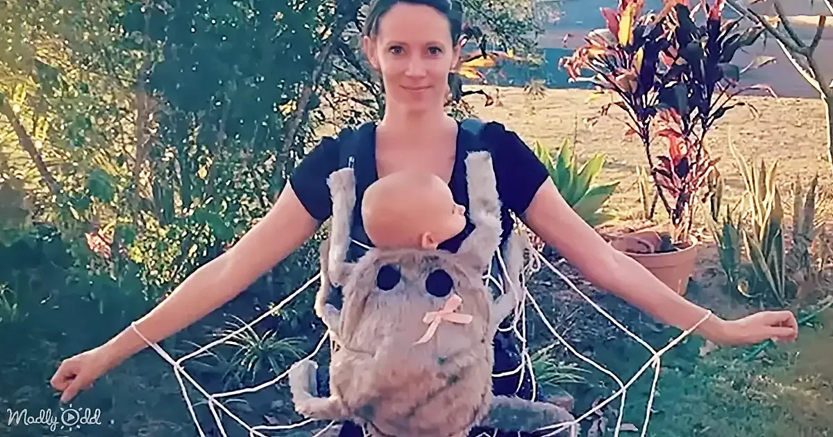 A woman in a blue dress holds a baby in a fur carrier, surrounded by greenery and a potted plant.