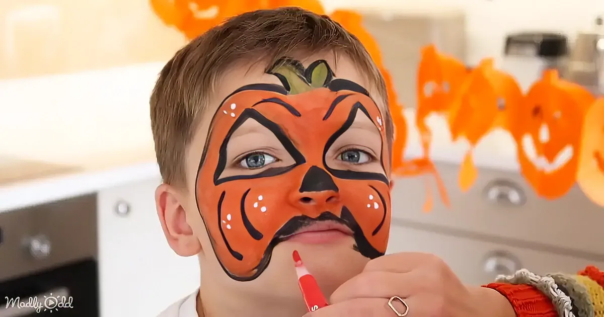 A child with a pumpkin face paint design, holding a red marker, in a kitchen setting.