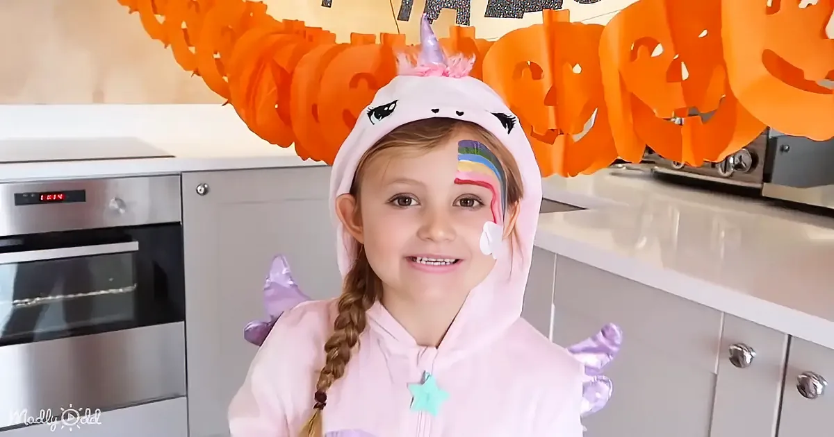 A girl in a unicorn hoodie smiles in a kitchen with Halloween decorations.