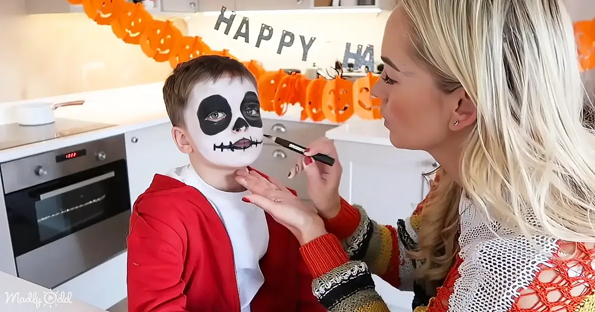 A woman applies skull makeup to a child's face in a kitchen decorated for Halloween.
