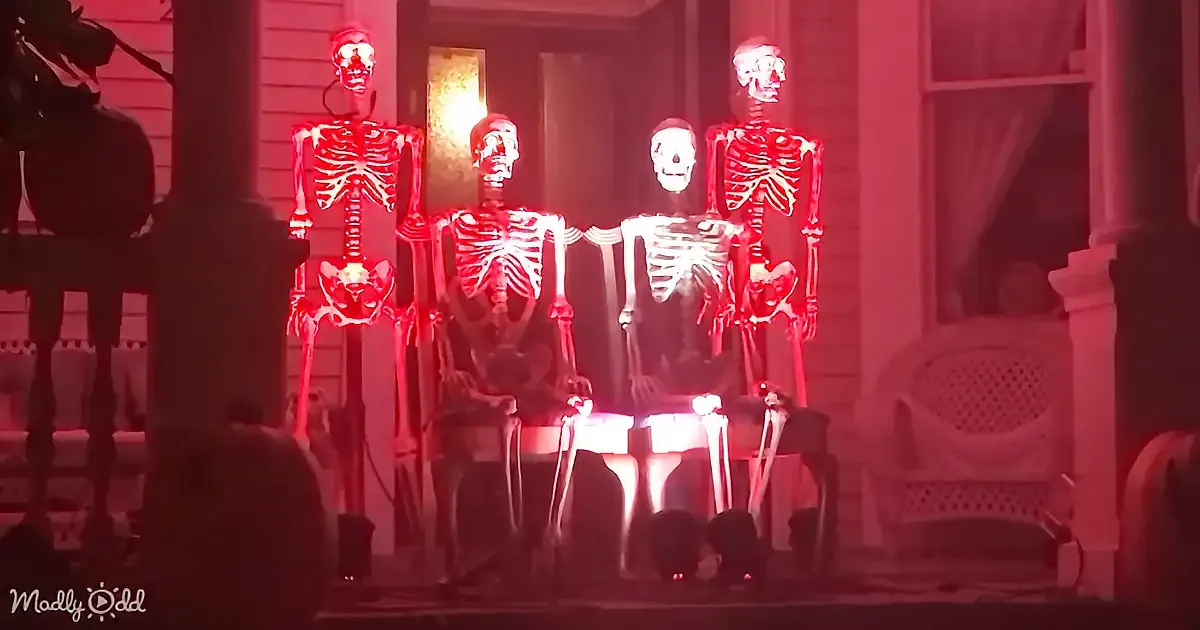 Four illuminated skeletons stand on a porch, bathed in red light, creating a spooky Halloween scene.