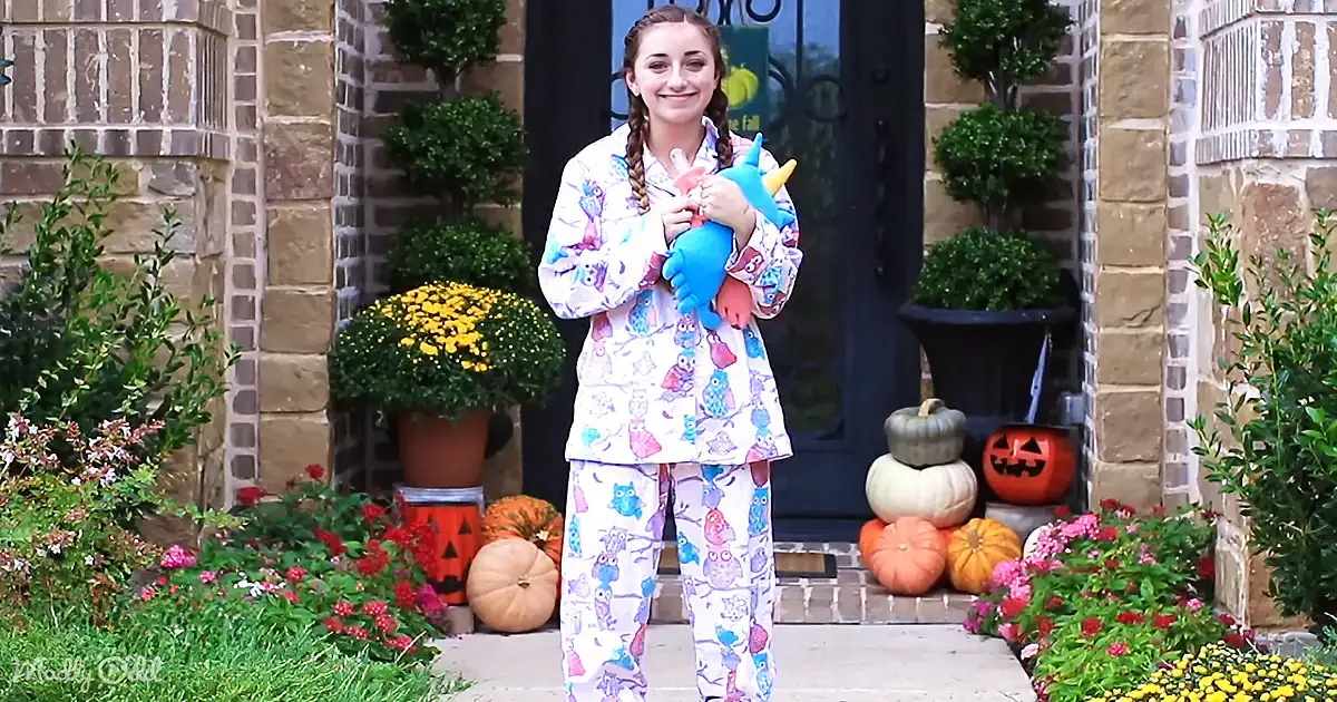 A girl in colorful owl pajamas holds a blue unicorn plush, standing in front of a door with autumn decorations.