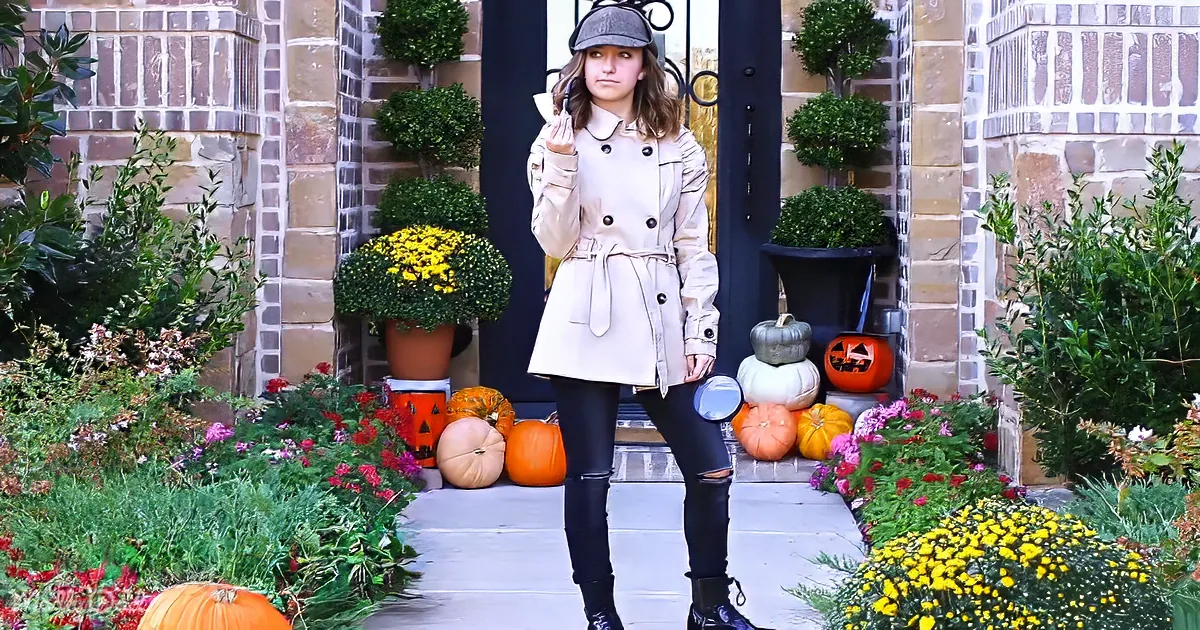 A woman in a beige coat and hat stands in front of a door adorned with pumpkins and flowers.