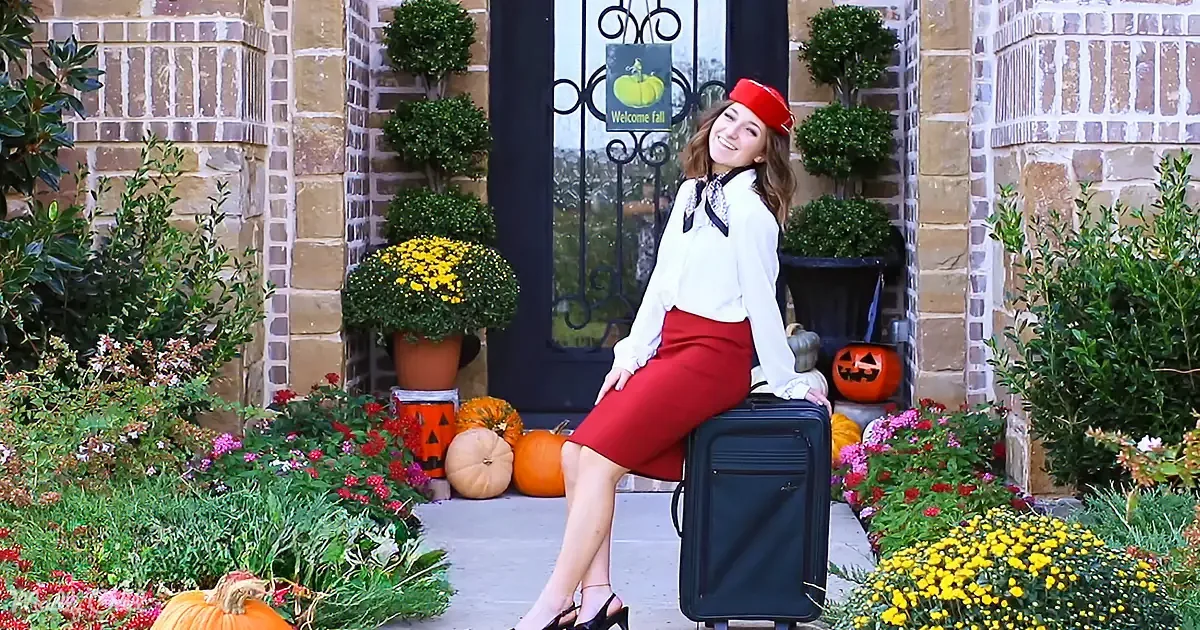 A woman in a red skirt and hat sits on a suitcase in front of a house decorated with pumpkins and flowers.