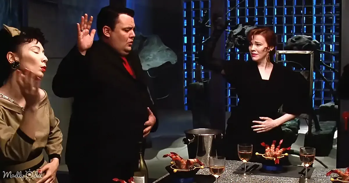 A man in a black coat gestures dramatically while two women react, with a lobster and wine glasses on the table.