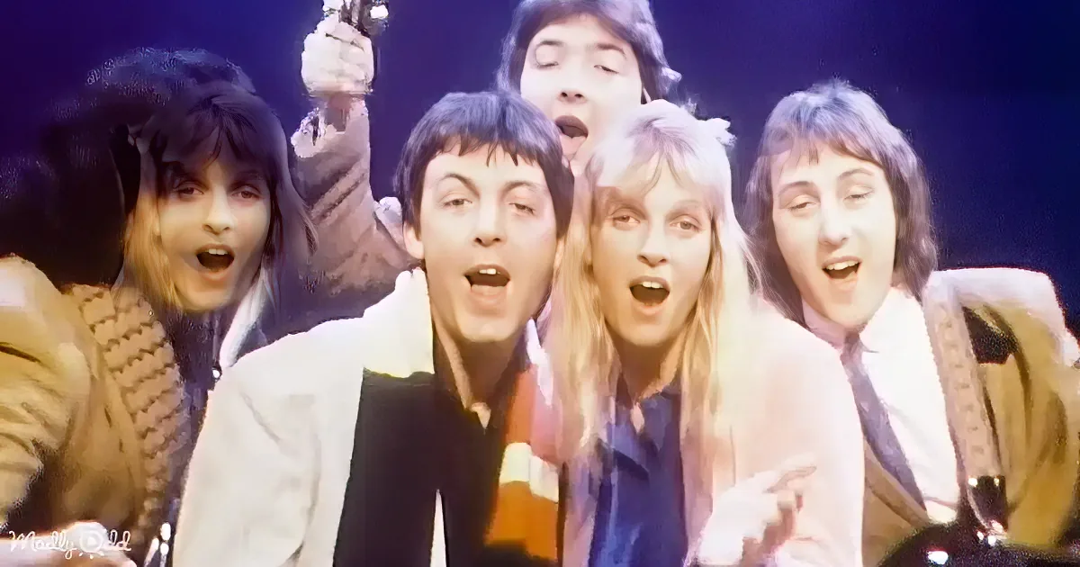 A group of people, including a man in a white jacket, are singing and smiling together on stage.