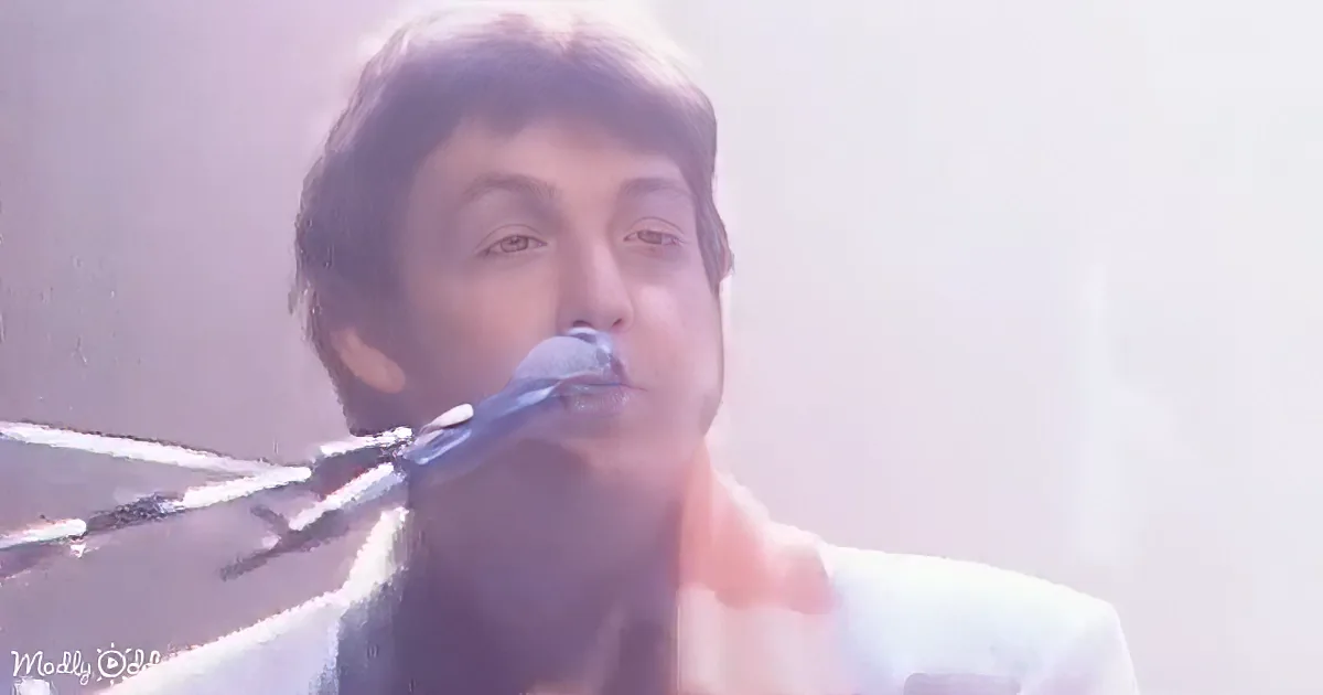 A person singing into a microphone, wearing a light-colored suit, with a blurred background.