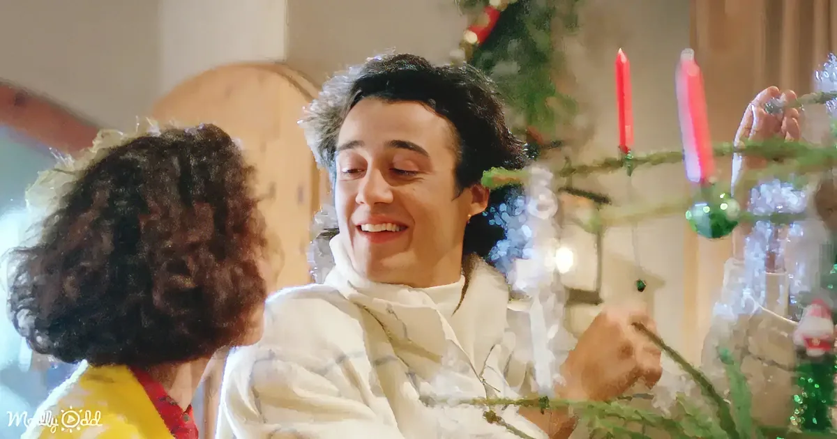 A man in a white shirt smiles while decorating a Christmas tree with a woman in a yellow top nearby.