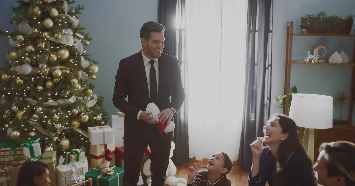 A man in a suit stands by a Christmas tree, holding a red object, while a family celebrates around him.