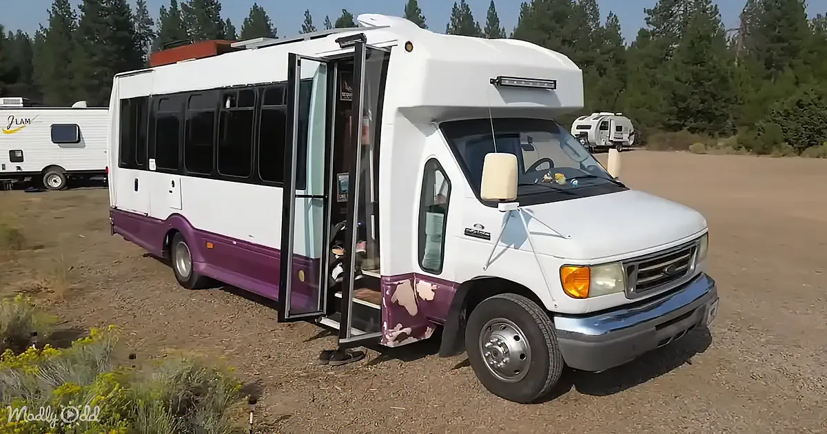 A white and purple RV with an open door is parked on a dirt lot surrounded by trees.