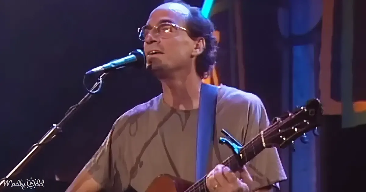 A man with glasses and dark hair is singing into a microphone, looking upwards.