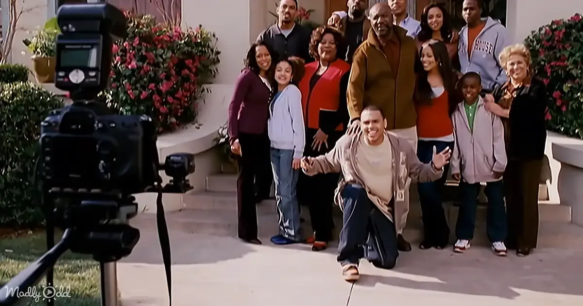 A man kneels in front of a group of people, smiling, as they pose for a photo in front of a house.
