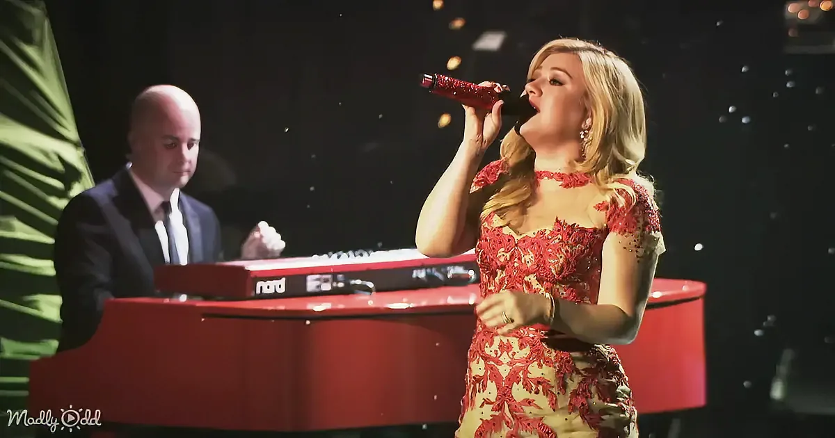 A woman in a red dress sings into a microphone while a man plays a red keyboard in the background.