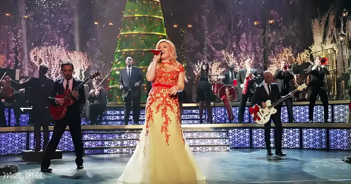 A woman in a yellow dress sings on stage with a band and a Christmas tree backdrop.
