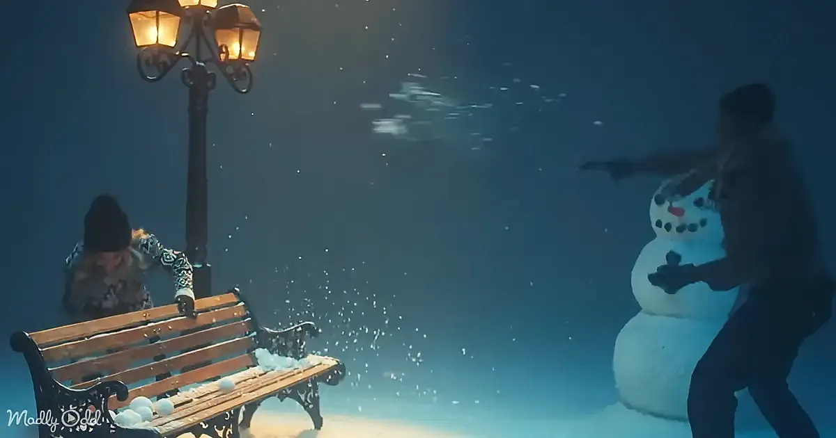 A person throws snowballs at a snowman while another person sits on a bench under a street lamp.