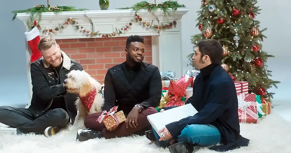 Three people sit near a Christmas tree, exchanging gifts and smiling, with a dog in a festive outfit.