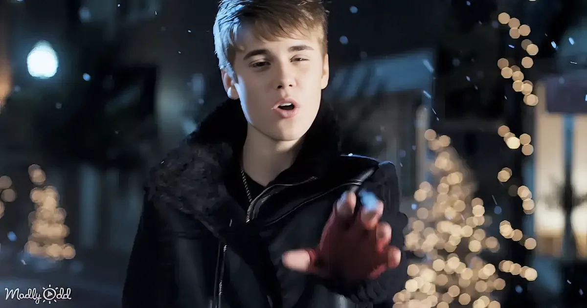 A smiling person in a black jacket stands under falling snowflakes, with blurred lights in the background.