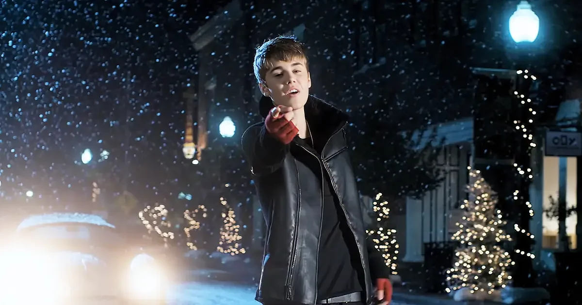 A person in a black leather jacket points forward under a snowy night sky, with Christmas lights in the background.