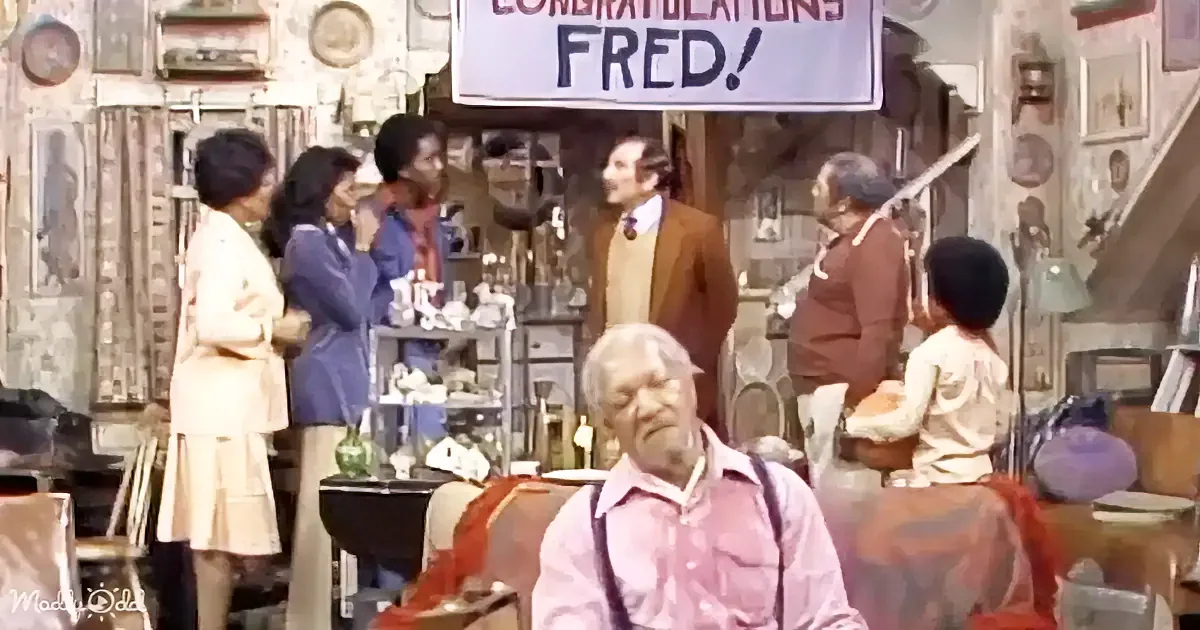 A man in a pink shirt sits in a cluttered room, surrounded by people and a sign saying "Congratulations Fred!"