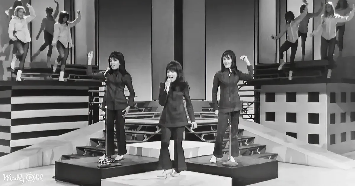 Three women in matching outfits sing on stage, with dancers in the background.