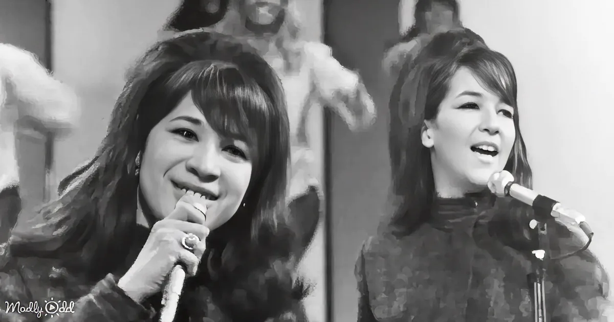 Two women sing into microphones, one smiling, both with long hair, in a black-and-white setting.
