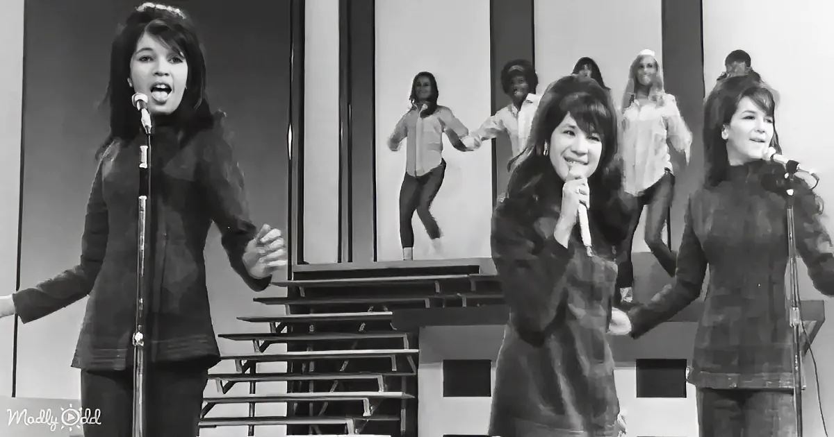 A woman in a black outfit sings into a microphone on stage, with a mirrored backdrop showing her dancing.