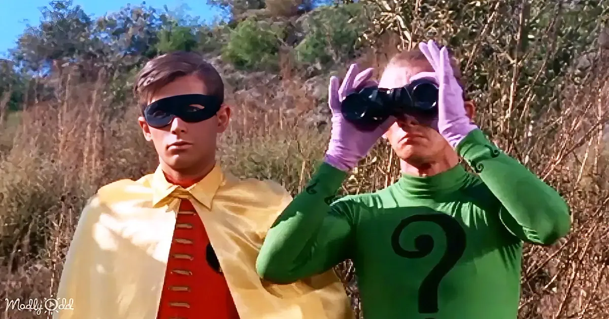 A man in a green Riddler costume with a question mark uses binoculars, while another in a yellow superhero outfit stands...