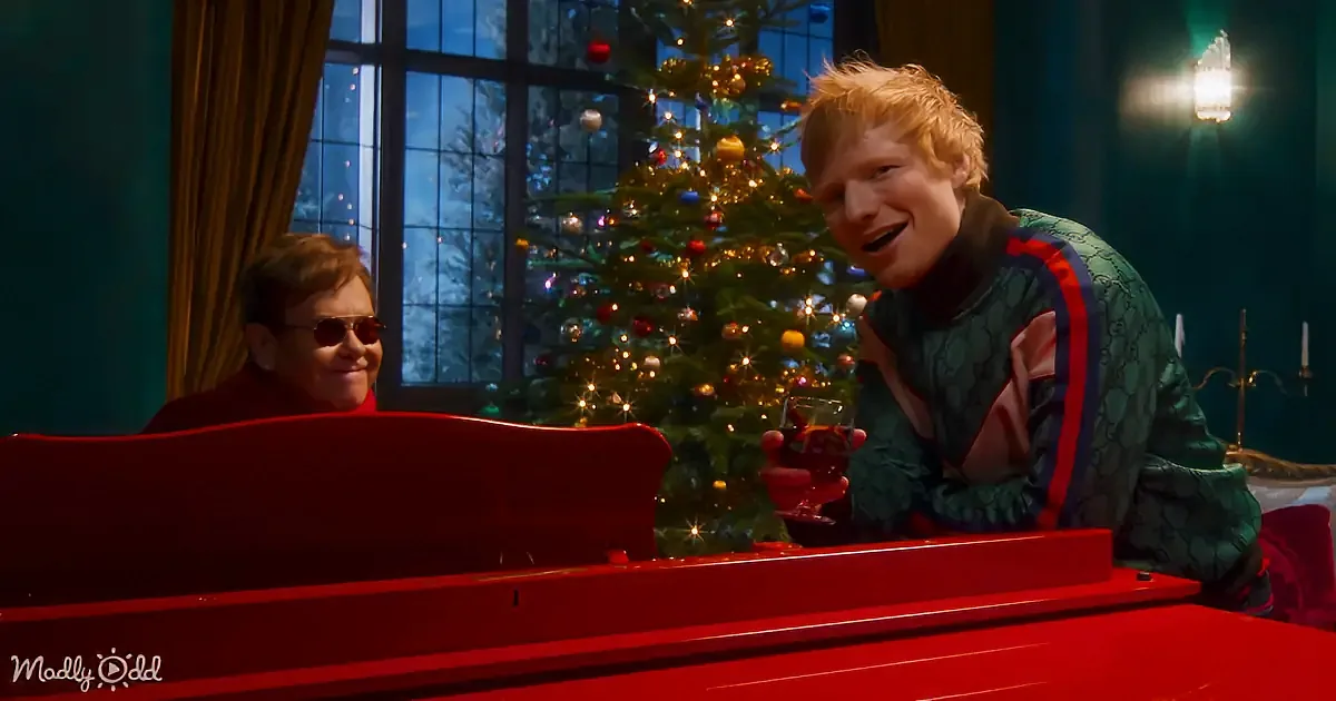 Ed Sheeran leans on a red piano, holding a drink, with Elton John in sunglasses nearby, both smiling near a Christmas tree.