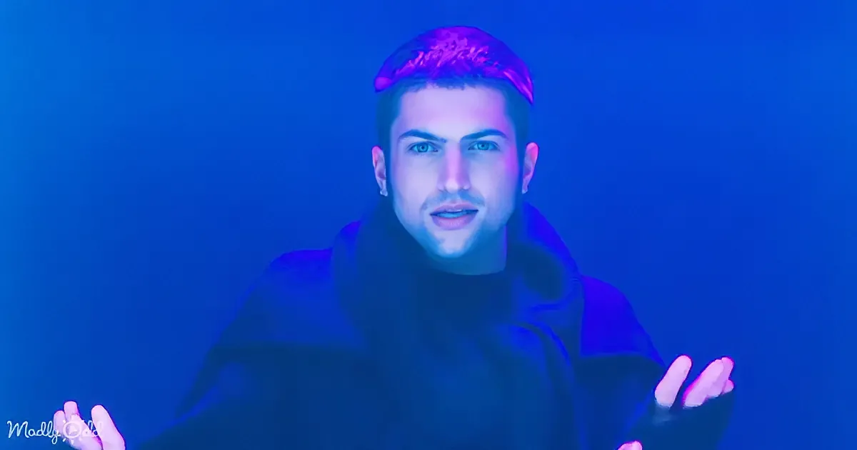 A person with short hair, wearing a dark hoodie, stands against a blue-lit background with their hands slightly raised.