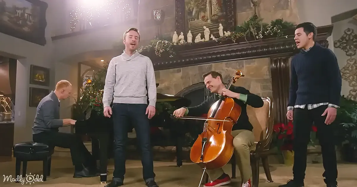 A man in a grey sweater sings while another plays cello, accompanied by a pianist and another singer in a cozy, decorated...