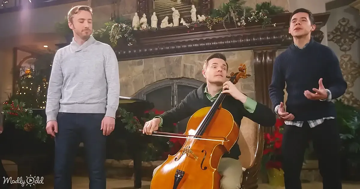 A man plays cello while two others sing in a festive, decorated room.