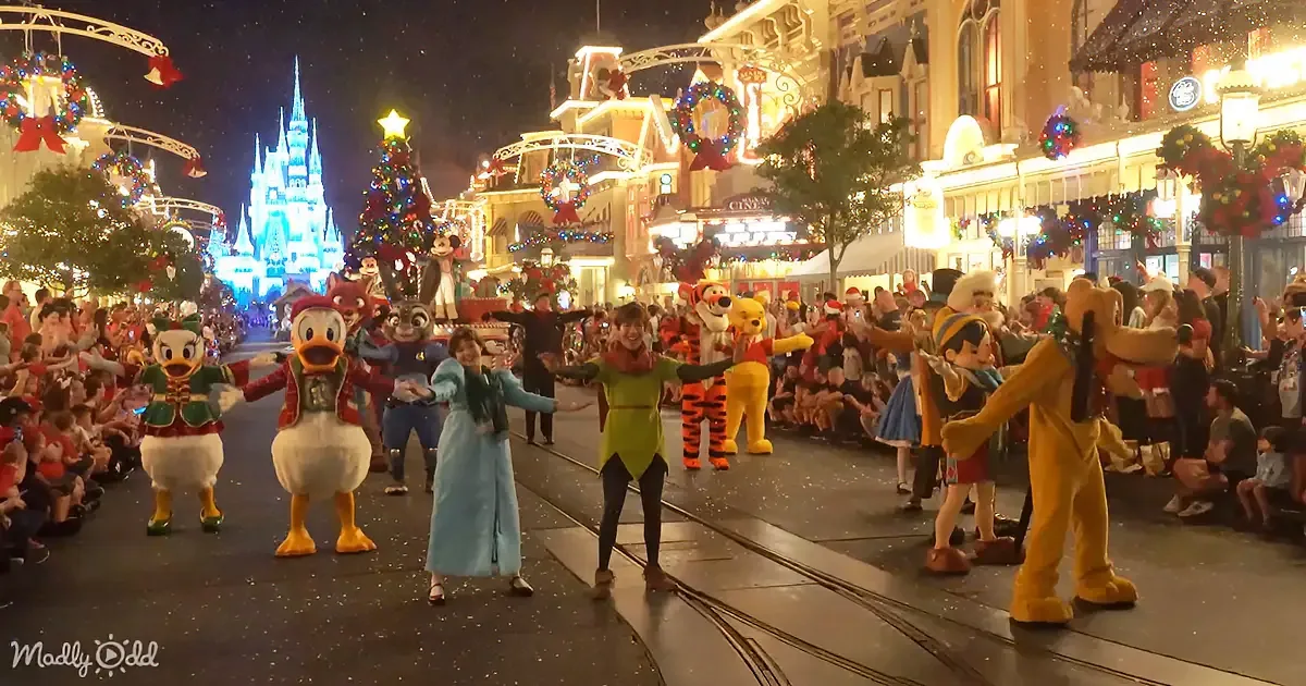Disney characters in festive costumes perform on a brightly lit street during a Christmas parade.