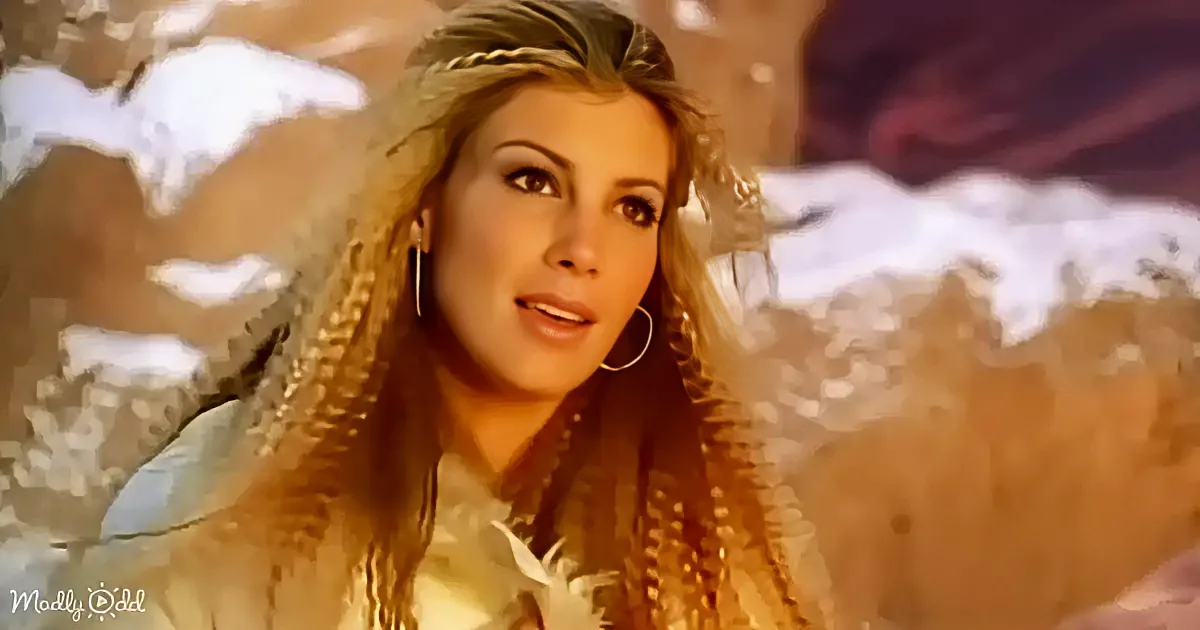A woman with long, braided hair and hoop earrings stands against a backdrop of mountains and clouds.