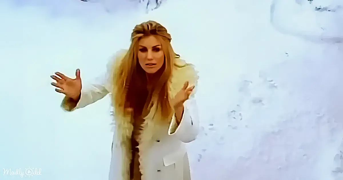 A woman in a white coat stands in the snow, arms outstretched, with long hair flowing.