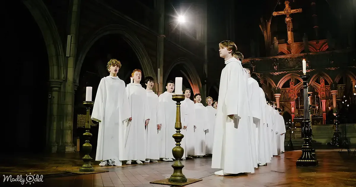 A group of young boys in white robes sings in a church, with a man in a white robe standing in the background.