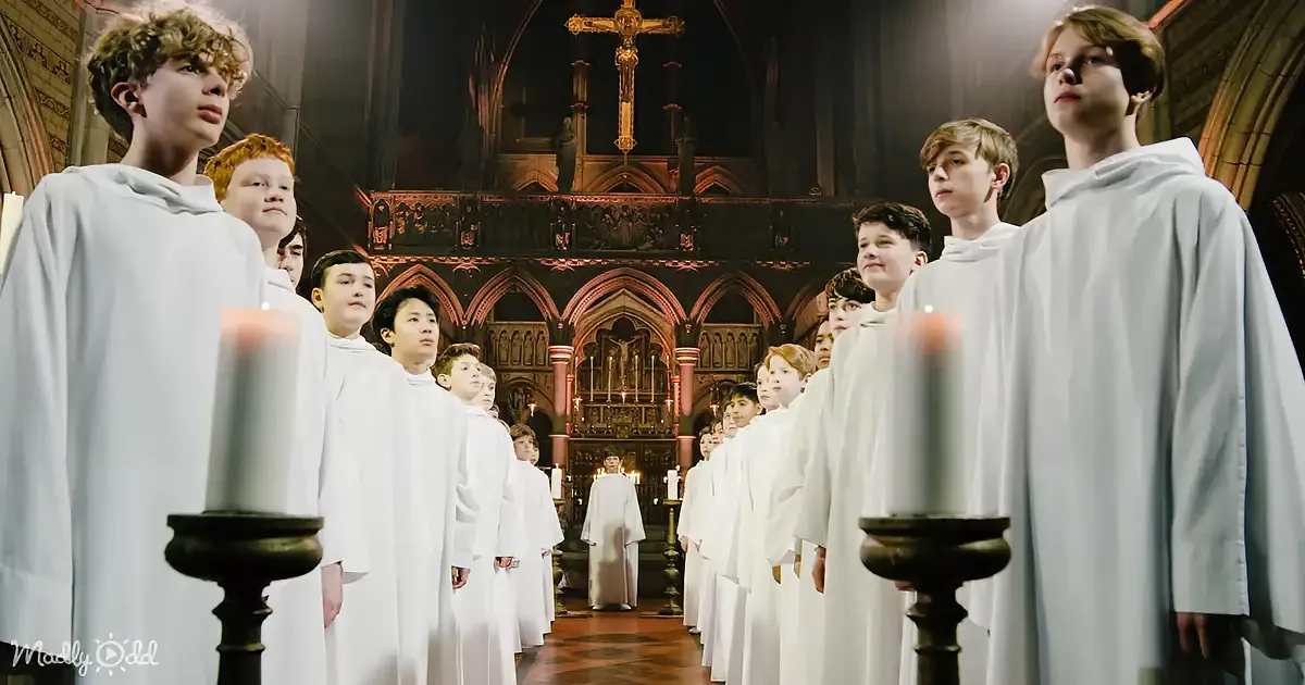 A group of boys in white robes stands in a church, holding candles, with a crucifix above.