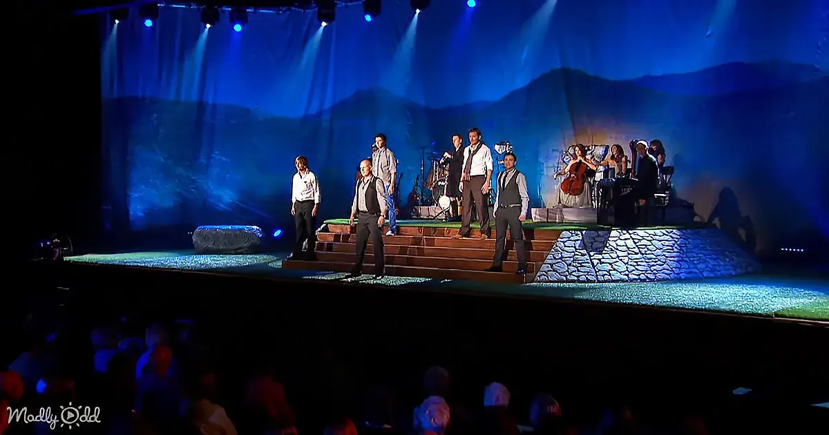 A group of men in formal attire stands on stage, accompanied by musicians, under blue stage lighting.