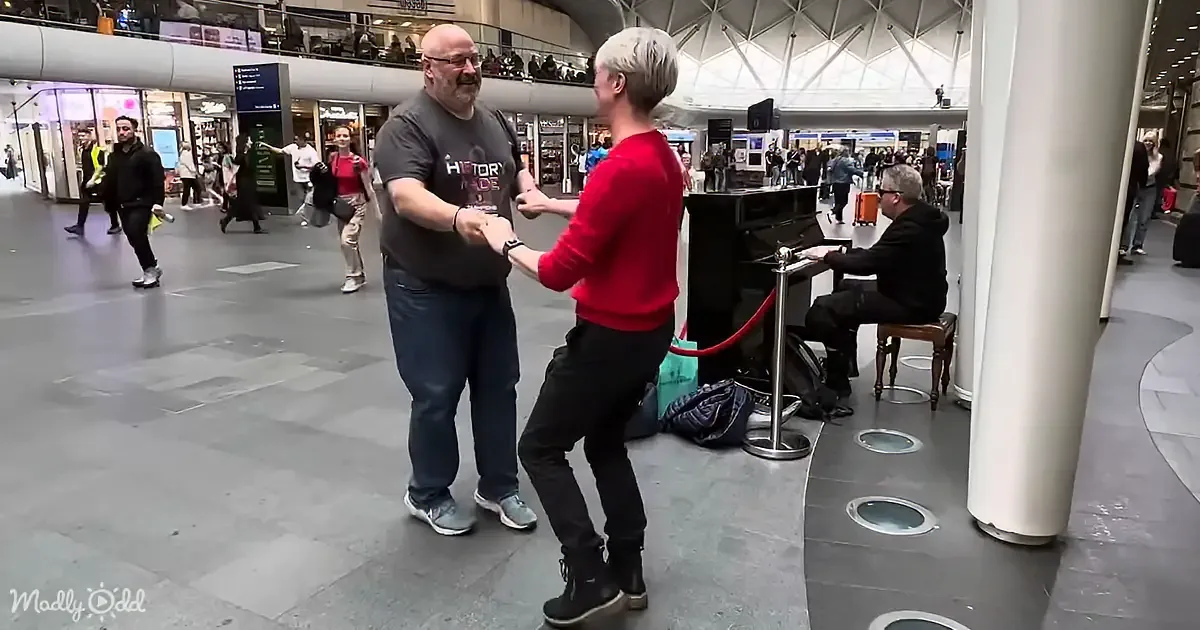 A man in a red sweater dances with another man in a black shirt at a busy airport terminal.