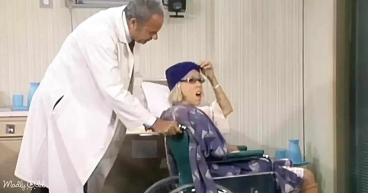 A doctor in a white coat leans over a woman in a wheelchair, who is wearing a blue headscarf and appears distressed.