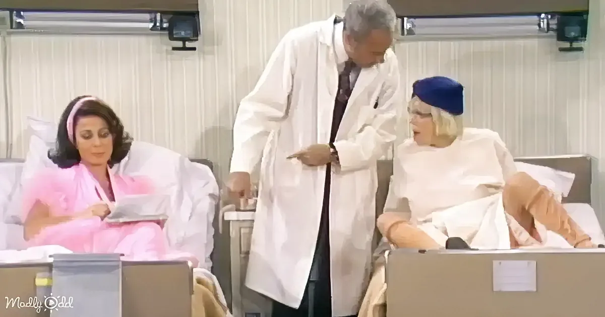 A doctor in a white coat stands between two women in hospital beds, one in pink and the other in white.